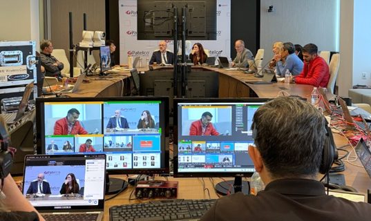 An annual meeting being broadcast from a boardroom, with cameras and monitors set up behind the table.