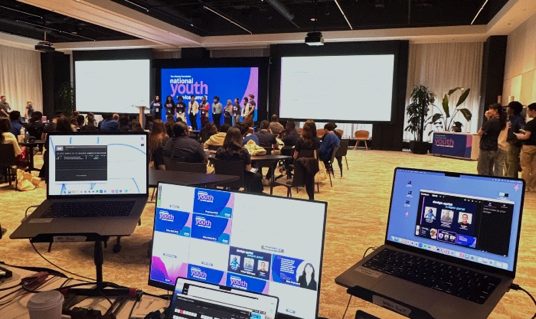 A conference general session in progress, viewed from the production position behind a row of laptops running the show.