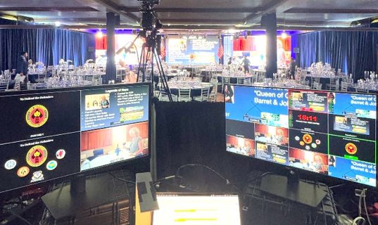 Production desk at a gala, monitors showing camera feeds and lower thirds over a ballroom of banquet tables.