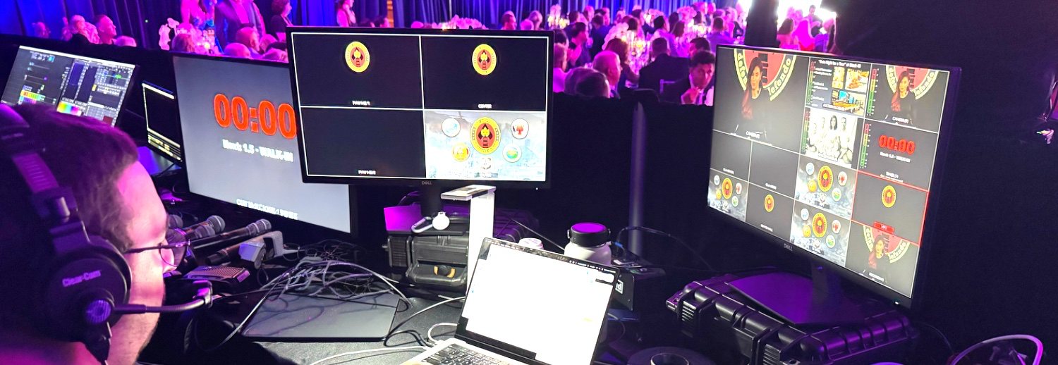 A show caller at the production desk during a gala, headset on, watching a countdown clock and a multiviewer of live camera feeds.